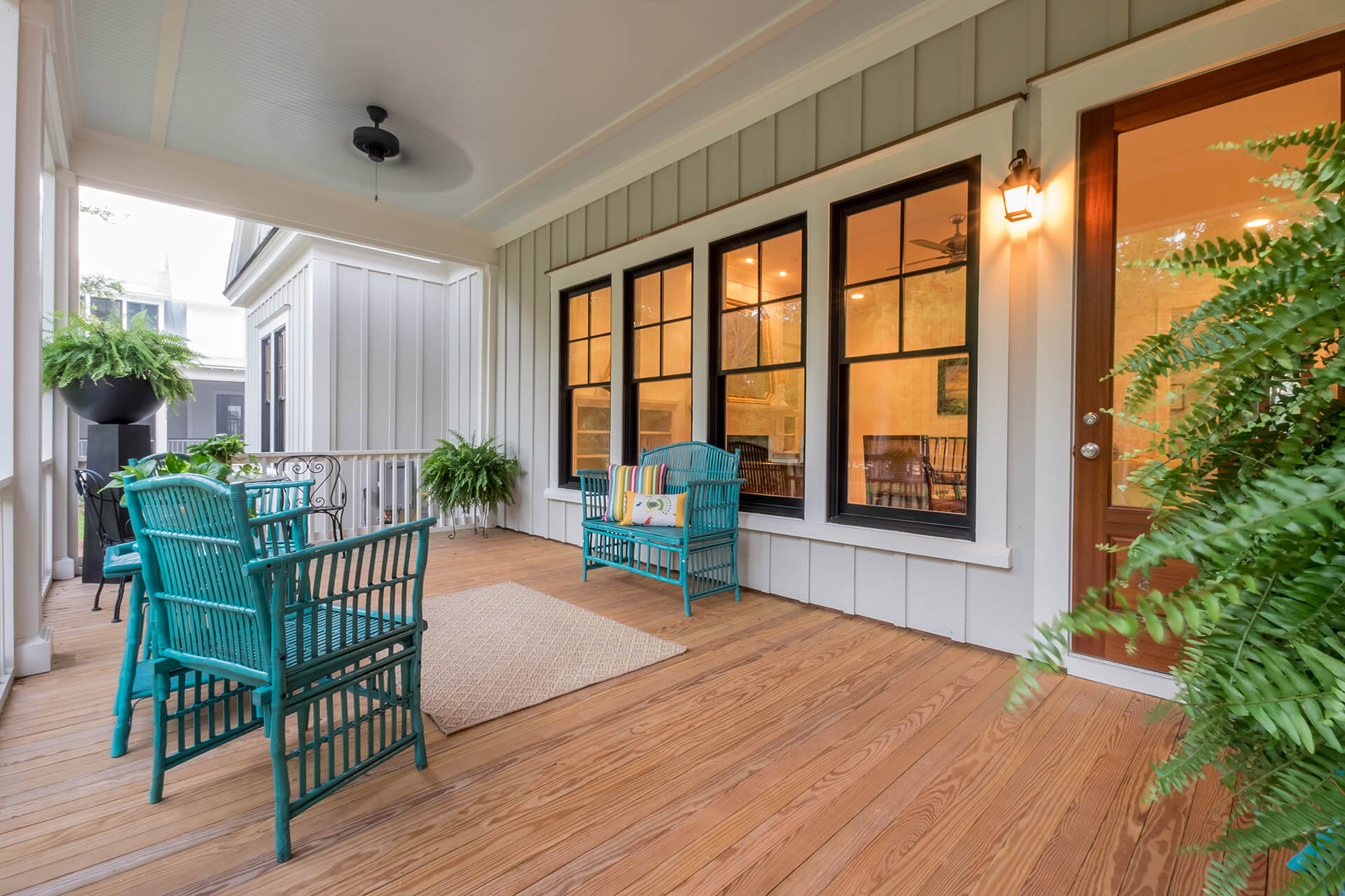 This porch has wood flooring with a tan rug in the middle. There are blue chairs and a blue bench for seating. There are various plants on the porch, and four windows show the inside of the house.