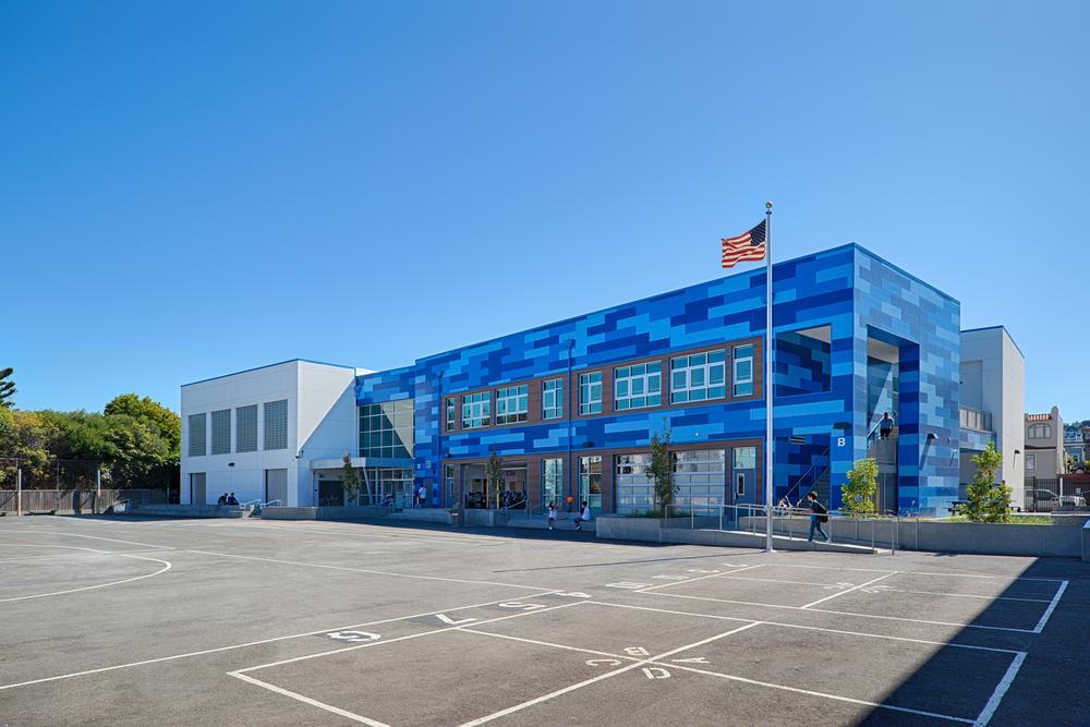 A far view of a bright blue school building, featuring three shades of blue from light to dark behind a flag pole flying the American flag.