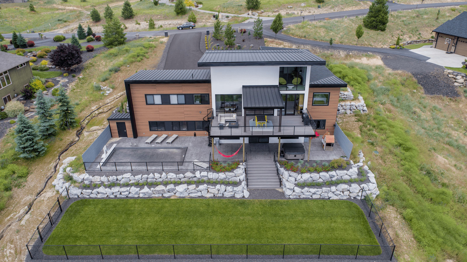 Aerial view of the back of a modern home, showcasing metal fencing, deck with spiral staircase, wood-textured exterior and patio sets.