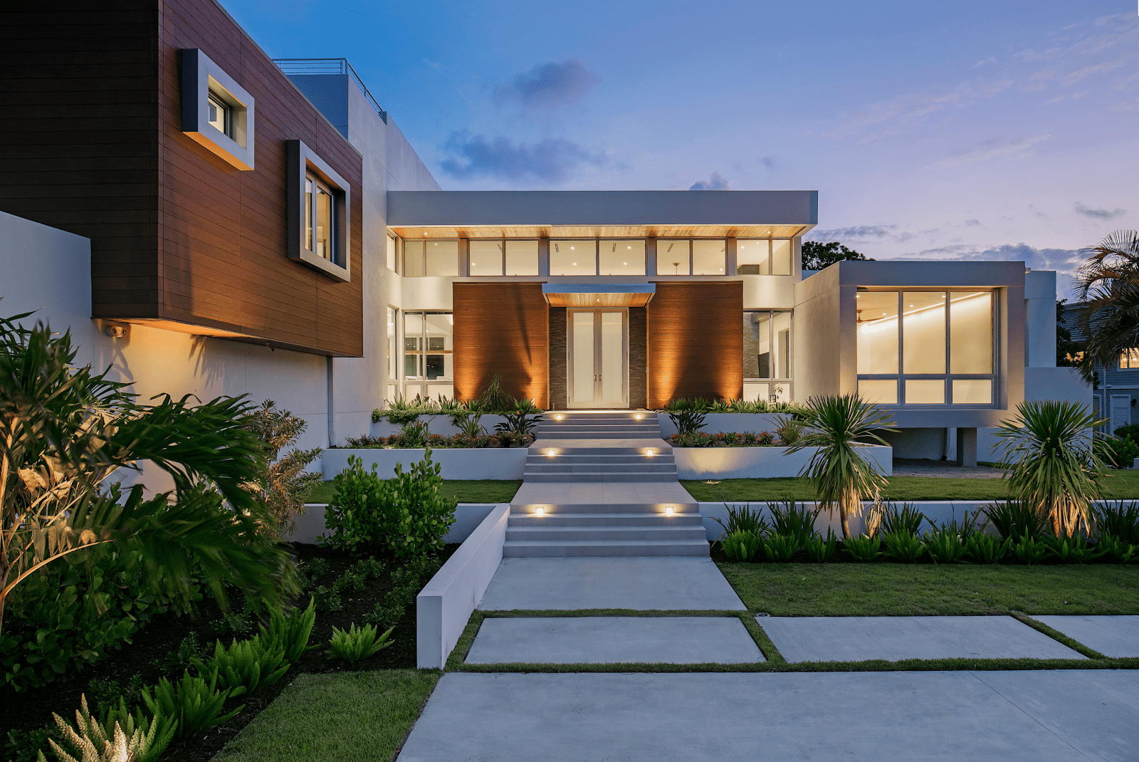 An exterior view of the front door at twilight, with spotlights on the pathway and in the side gardens. 