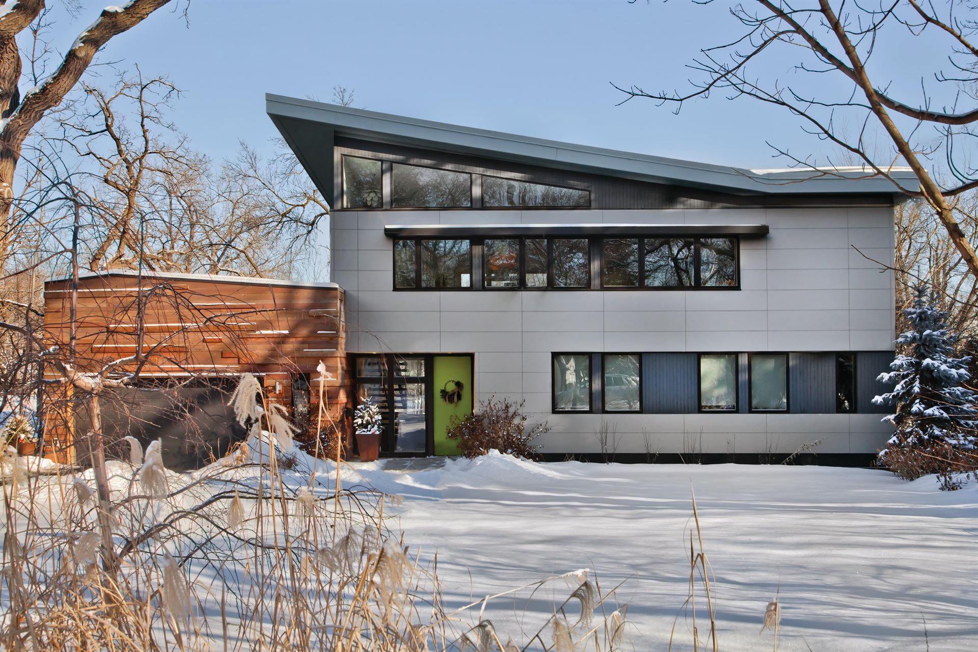 A snowy view at the front of a modern home, featuring gray Architectural Block siding and an upwards tilted roof.