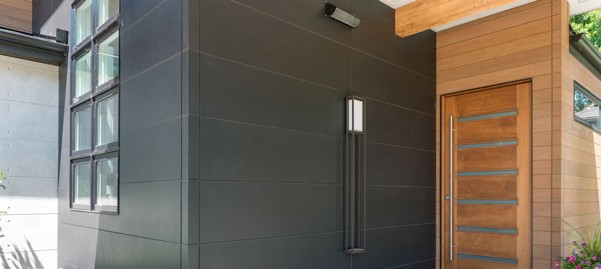 A close-up view of the entrance of a modern home, with a light wood-textured front door next to wide black siding panels.