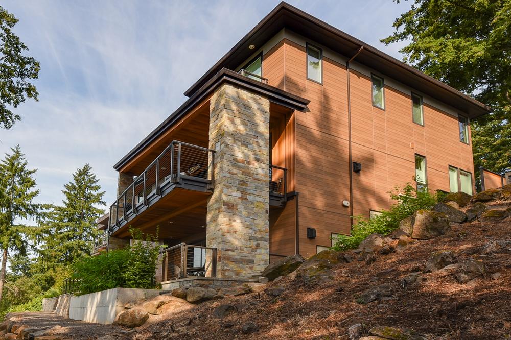 Home with light brown siding and stone column sits on a slope surrounded by pine trees.