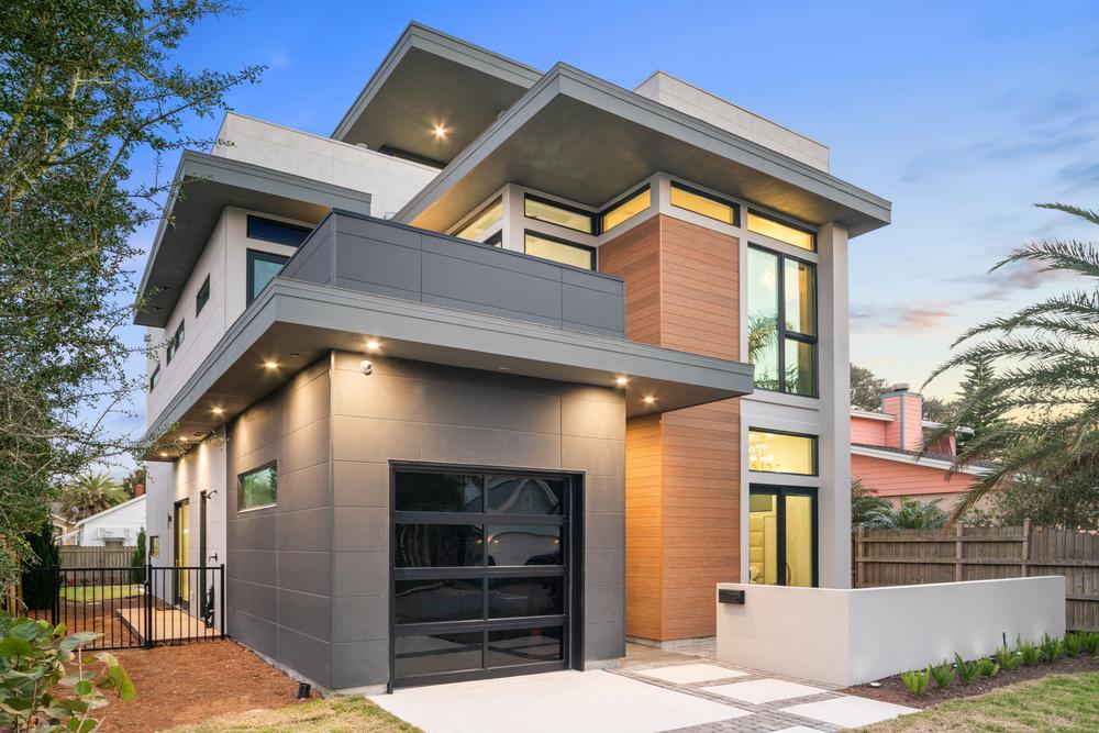 A modern, three-story home with a wooden pillar, glass garage door with black trim and gray siding.