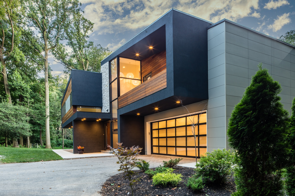 A black, brown, and gray home covered in Nichiha Fiber Cement Siding sits. Their lines are square and sharp, with bushes in the lower right corner, a driveway leading up to a paneled garage door, and a yard full of trees in the back left corner.