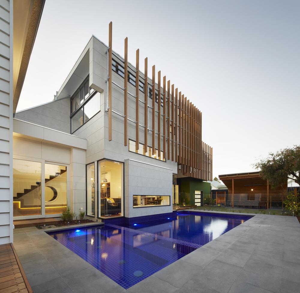 An off-white home with wooden planks on the exterior as side decor is shown from the back yard. There is a dark blue pool and the home is decorated with wide windows looking in, where there’s a staircase and an office-like room.