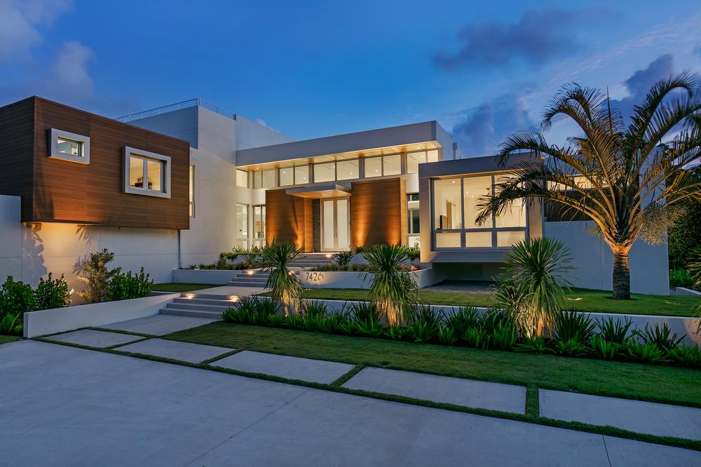 A white and brown home utilizing Nichiha Fiber Cement Siding sits at dusk, with palm trees in the front yard and lights illuminating it against the purplish-blue sky.