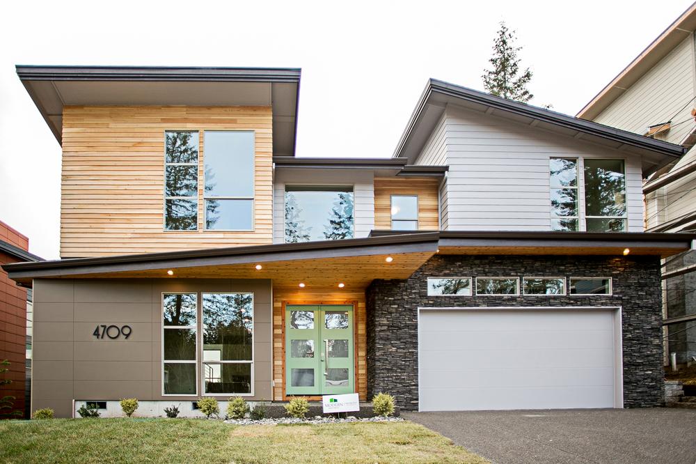 A modern home has an attached garage. The front, French doors are green, and there are various sidings incorporated into the design.