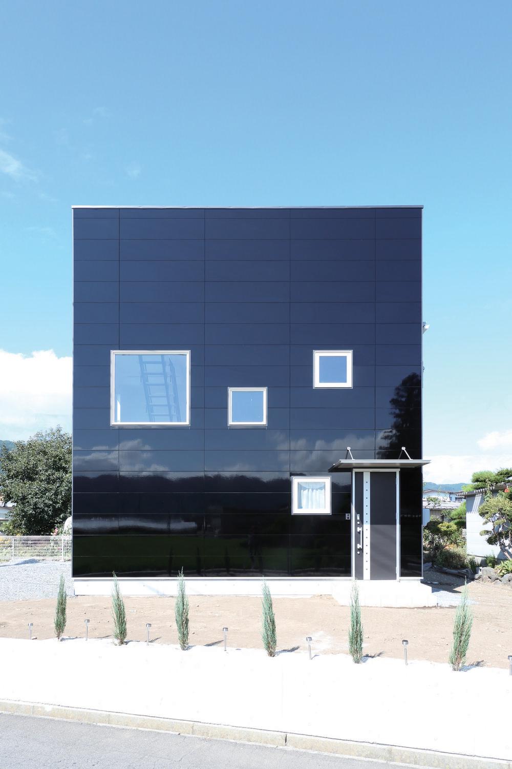 A glossy, black home in Nichiha Miraia siding is the focal point of this image. The house has a door on the right and four differently-sized square windows. There’s a blue sky and a few thin, tiny bush trees in front of the house by the sidewalk.