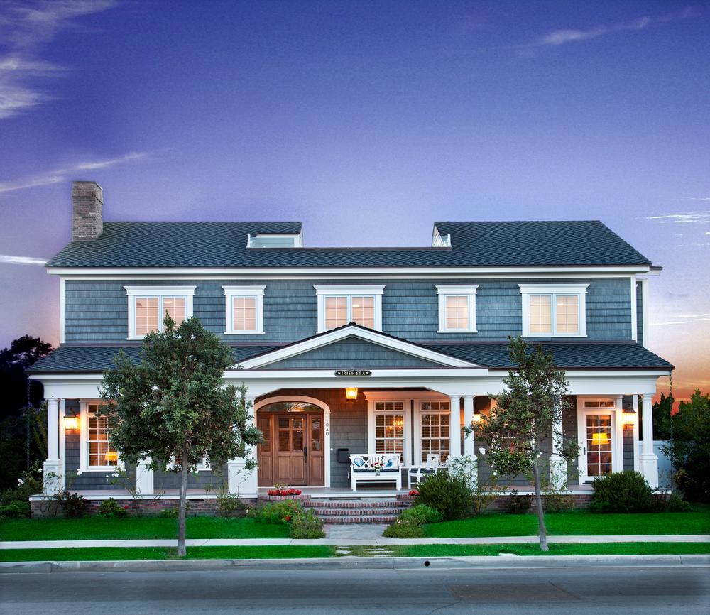 Rendering of home with blue cedar shake look fiber cement paneling. There is a sunset in the background and the porch and window lights are on in the home.