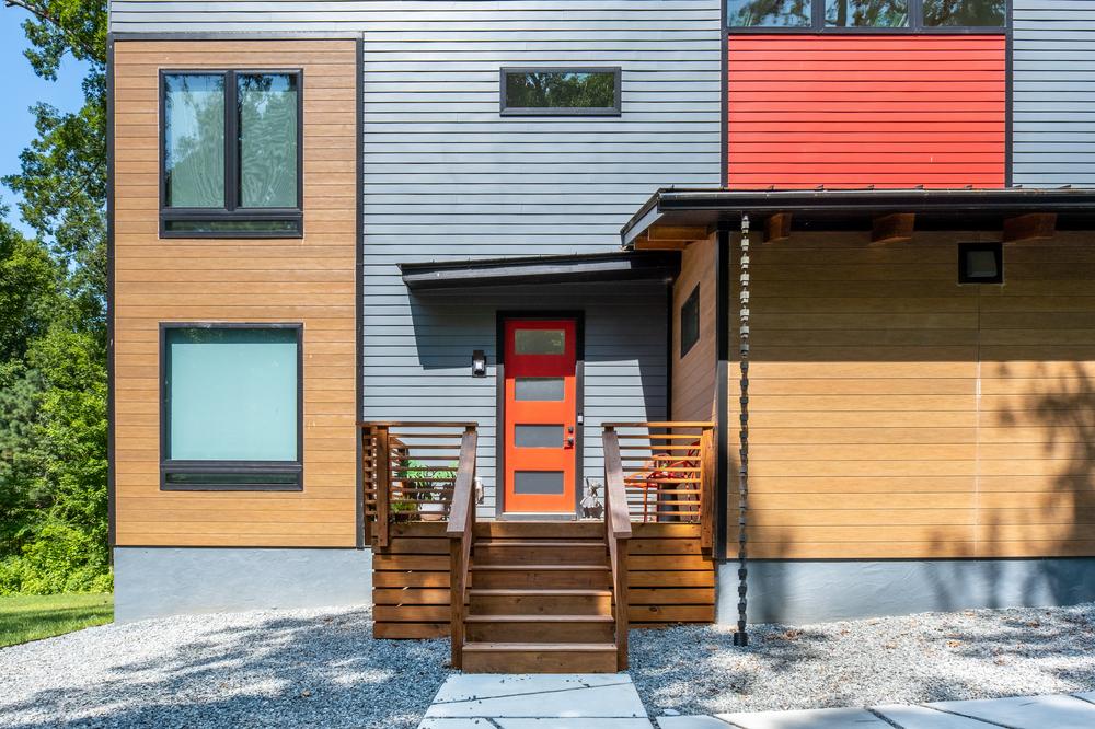 A home has grey, red and wood-like siding. There is an orange front door and a small patio with a plant and orange chairs on it.