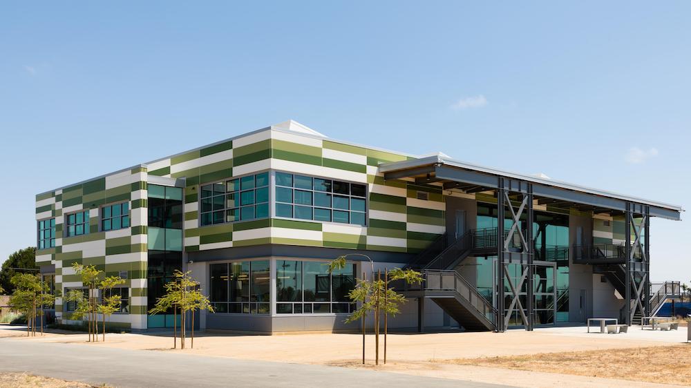 Mount Eden High School, a two-story building clad in Nichiha’s Illumination panels in white, light green and dark green is standing in front of a cement walkway and a few small trees.