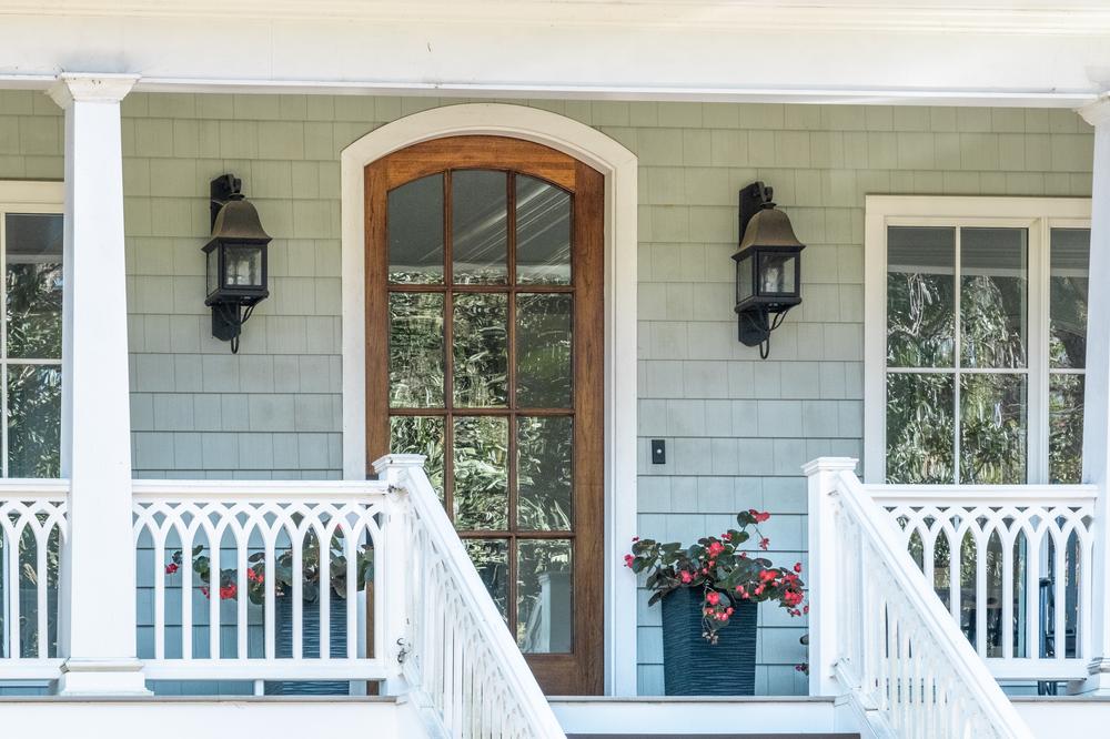 This close-up image shows a home&rsquo;s front porch. There are fiber cement cedar shakes in a light blue color. There are two lights on the sides of the wooden front door, and there are two flower pots below the lights.