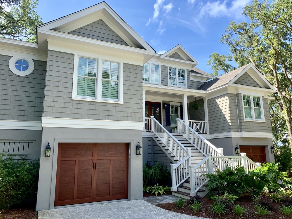 This three-story house has two garages with wooden doors. There is a large stairway leading to the front door. The siding on the house is fiber cement cedar shake in a light grey.