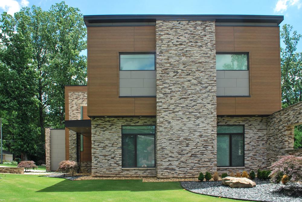 This two-story has a brick, wood-like siding with gray cement touches. There is a green yard on the side of the home.