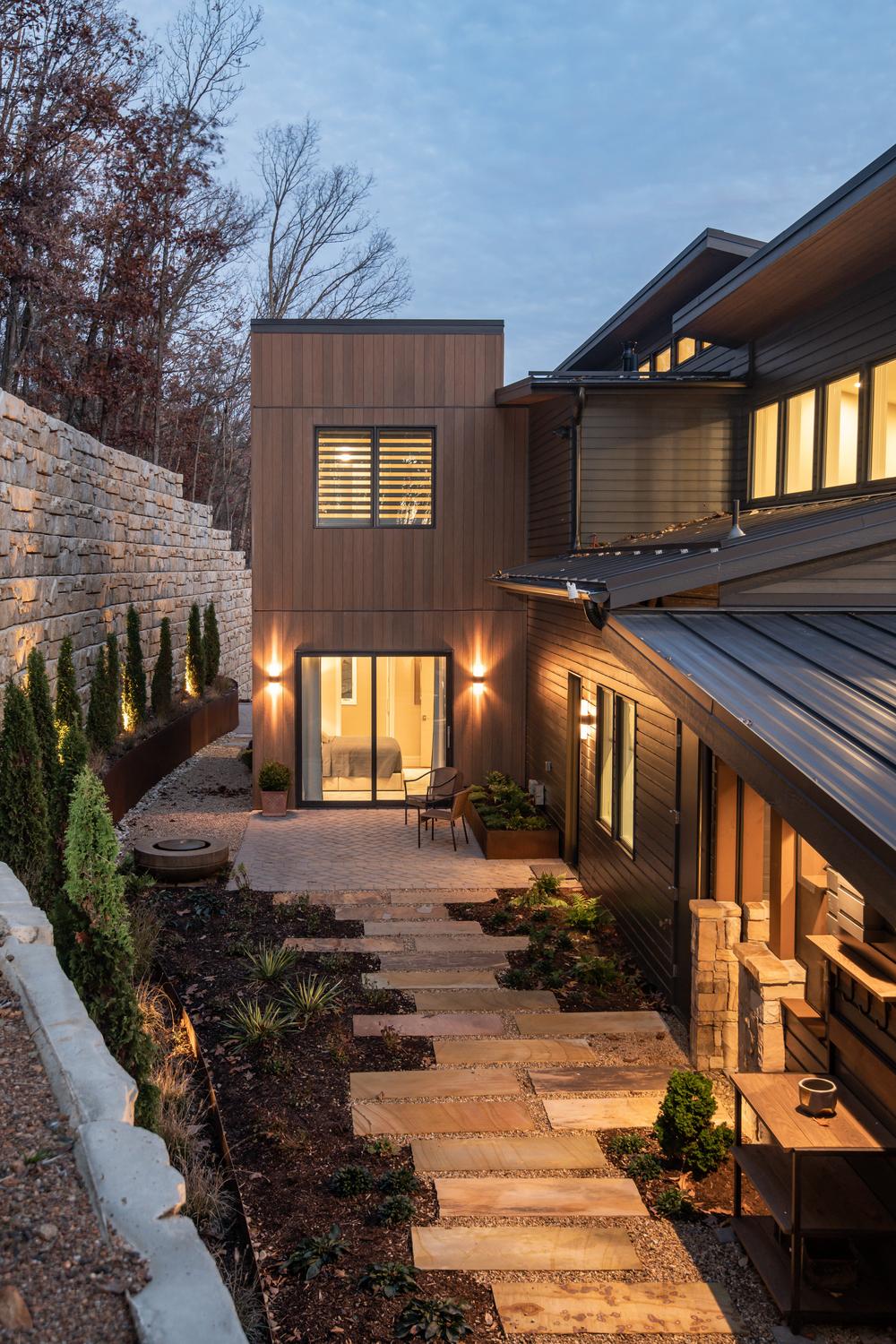 The back of this home has a small patio area. The home has a wood-like siding, and there is a stone wall that separates the woods from the residence.