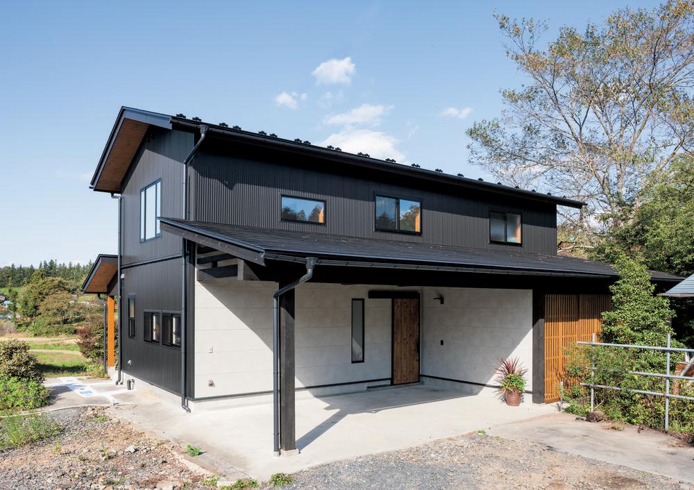 A black home decorated in Nichiha’s fiber cement siding with brown under trim, a black roof, and a white carport wall stands in the sun. There are trees in the background and a blue sky.