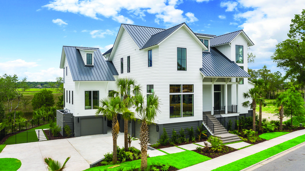 An off-white home decorated in Nichiha Fiber Cement Siding is the focal point. It has dark grey roofing, tall windows, steps leading up to a porch, and a lower-level garage under the home with dark siding. The lawn is full of palm trees, shrubbery, and there is blue sky with white clouds behind it.