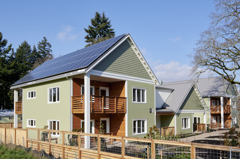 A mint green home with brown accents and a solar panel roof sits in the sun with a backyard garden and trees in the background.