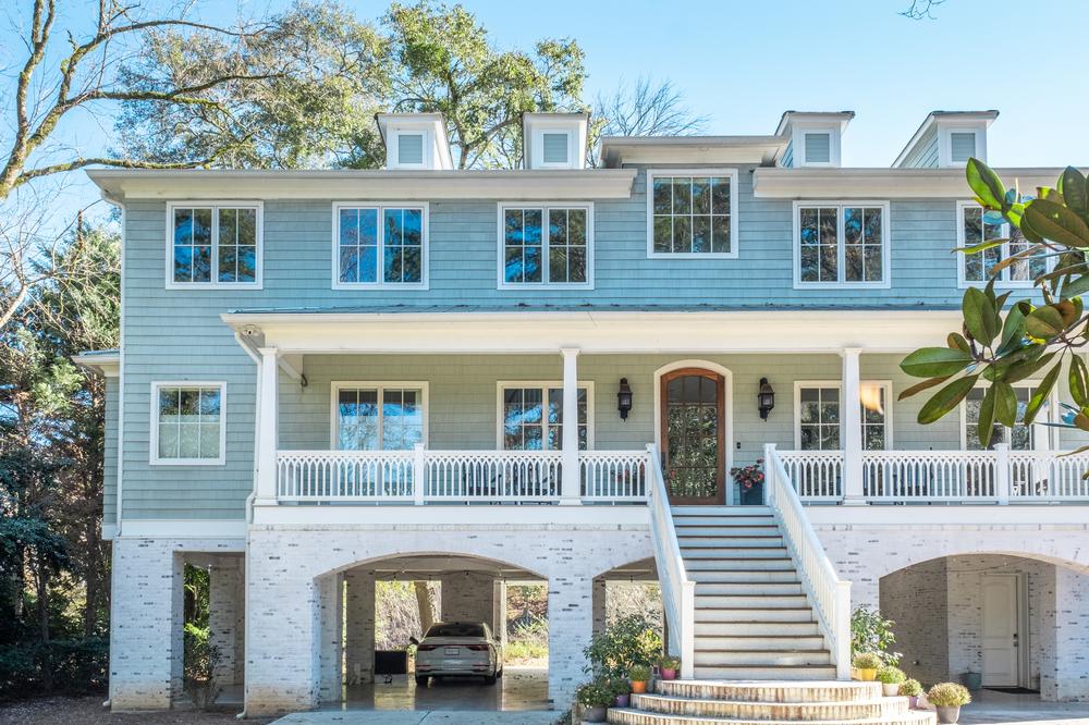 A light blue house decorated with Nichiha fiber cement siding with stone pillars underneath and an open garage. There are trees behind the home and on the right side. The windows are square with white trim and there are long steps leading up to the brown, glass front door.