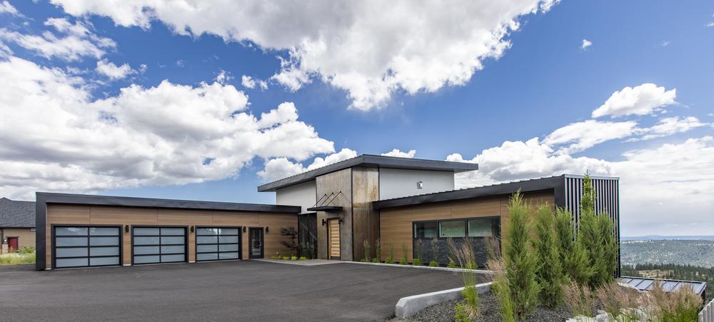 A brown, one-level home with brown siding, big garage doors, a black roof, and an elevated floor in the middle sits atop a hill with shrubs on the left, an open driveway, and a blue sky with white clouds behind it.