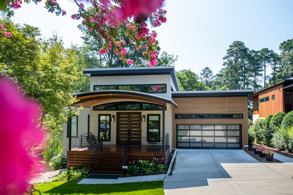 A home in Raleigh with Nichiha fiber cement siding is centered underneath a tree with pink flower blossoms. The home has a driveway on the right with a brown garage door add-on, white siding, and a front porch with a thin roof.
