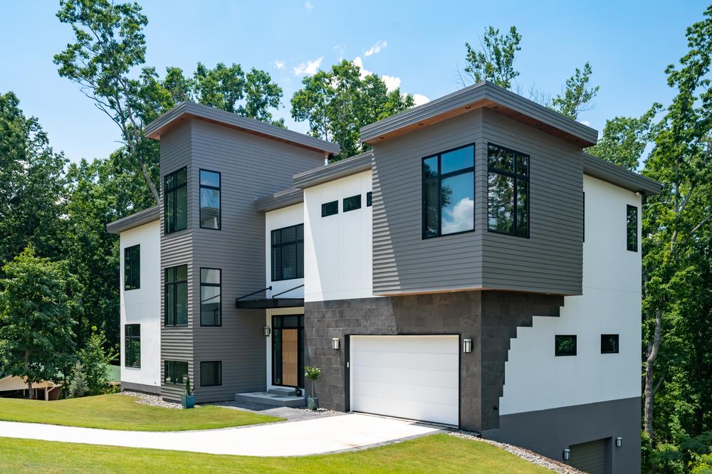 A grey, stone, and white accented home with Nichiha’s fiber cement siding is surrounded by a green lawn, a blue sky and a plethora of green trees.