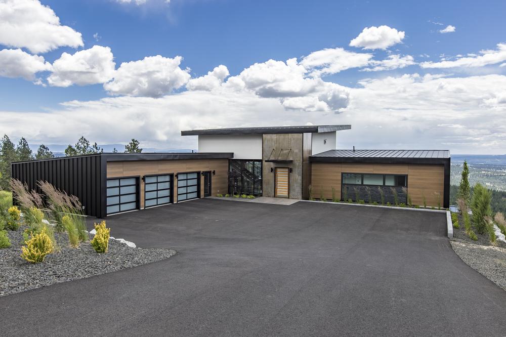 A brown, black, white, and stone accented home protected with Nichiha’s fiber cement siding sits atop a hill with a large driveway and blue, cloudy skies in the background.