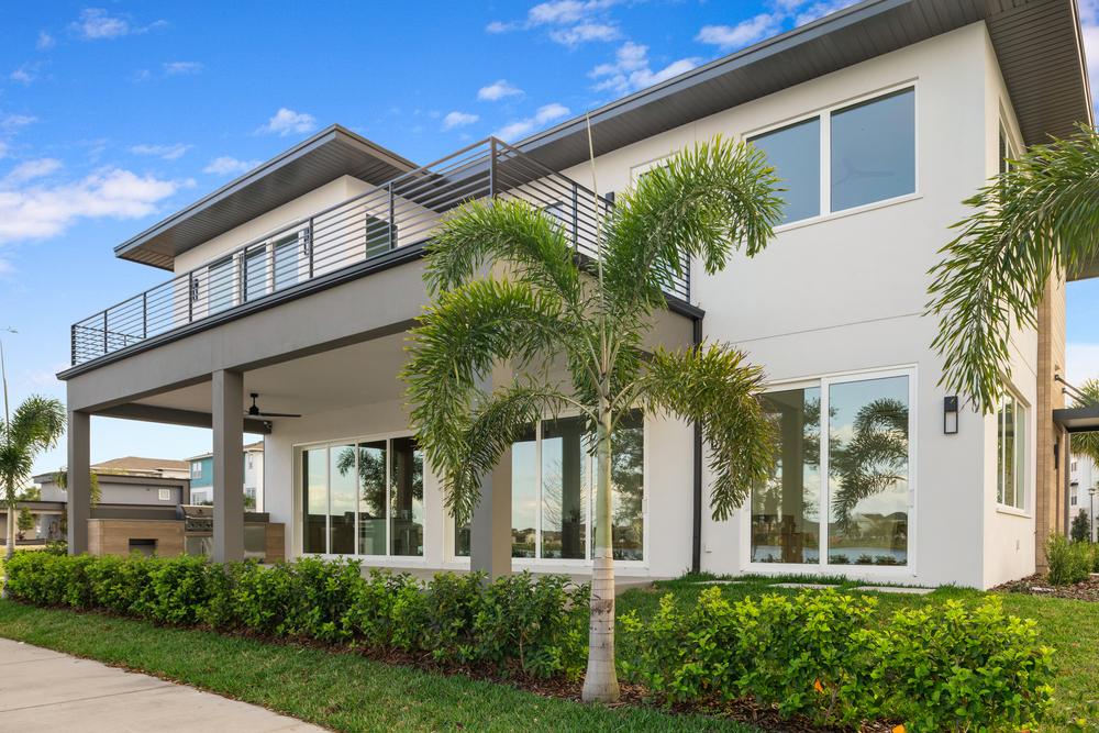 This modern Florida home has large windows on both stories. There is green grass surrounding the home with palm trees scattered throughout the landscaping.