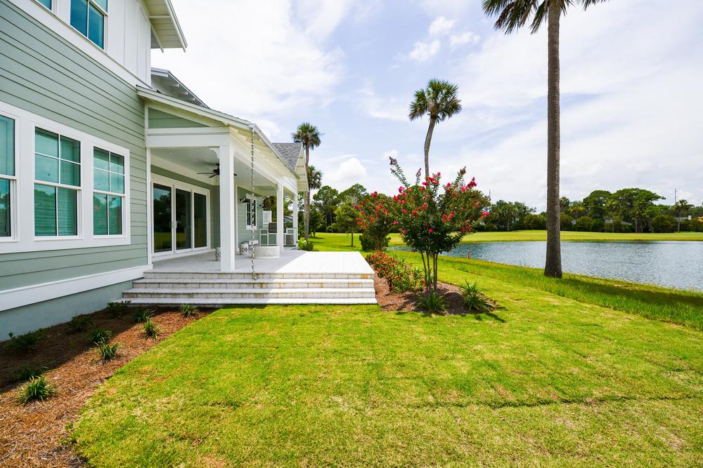 A home with blue colored siding with a big, white back porch overlooking the water with palm trees and other shrubs off the porch.