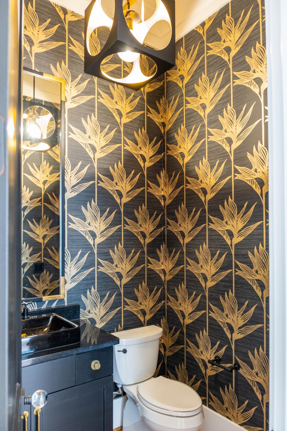 A bathroom with gold and black wallpaper in a floral pattern, a white toilet and black sink countertop. 