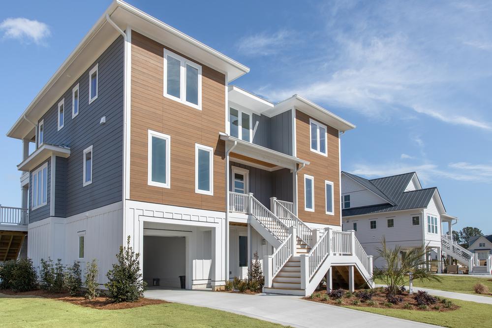Three-story home with brown and gray horizontal siding and tropical plants in the yard.